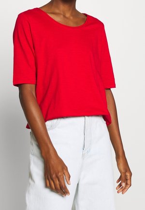 Osoba ubrana w jasnoczerwoną koszulkę z krótkim rękawem i jasnoniebieskie spodnie z wysokim stanem, przedstawiona od szyi do połowy uda na jednolitym tle.