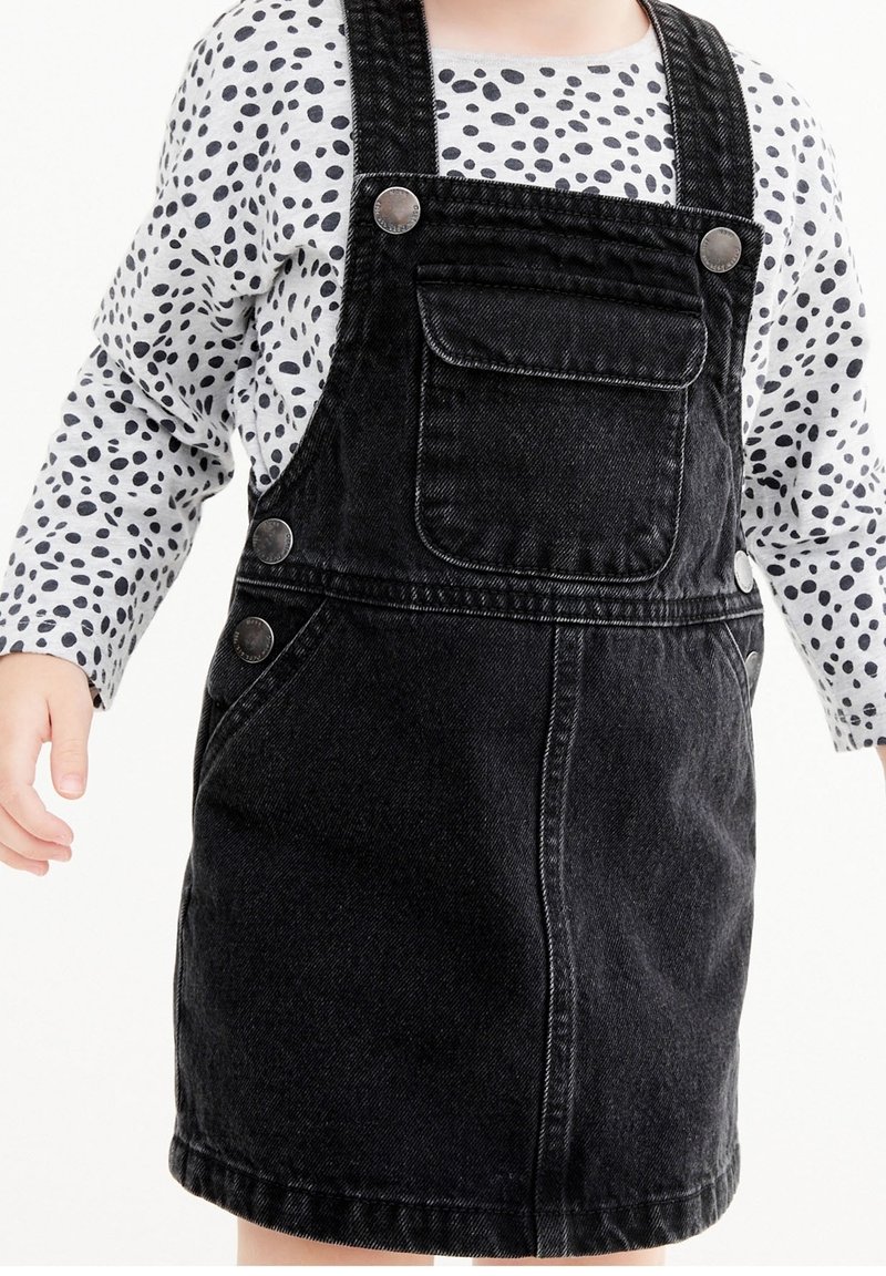 Enfant portant une robe salopette en denim noir avec poche avant et une chemise blanche à manches longues avec des taches noires, debout devant un fond blanc.