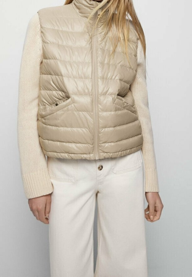 Beige puffer vest with horizontal quilted design, high collar, and side pockets, layered over a cream knit sweater. Neutral color palette.