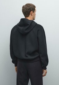 Man in een zwarte hoodie en donkere broeken, staand tegenover een lichtgrijze muur met ontspannen handen langs de zijkanten.