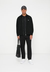 Juodas tekstūruotas užtraukiamas džemperis, juoda marškinėliai, juodos kelnės, baltos sportbačiai, pilka kepurė su logotipu ir juoda rankinė.