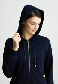 Felpa con zip di colore blu navy, dotata di cappuccio, con accessori in tonalità oro, lacci e due tasche laterali con bottoni decorativi. Texture liscia.
