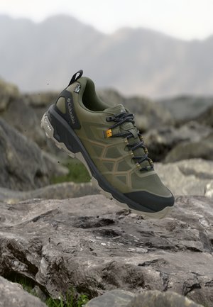 Olivgrüner und schwarzer Columbia Wanderschuh mit schwarzen Schnürsenkeln, der über felsigem Gelände schwebt, mit verschwommenen Bergen im Hintergrund.