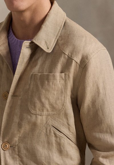 Beige herringbone jacket with two front pockets and wooden buttons worn over a lavender knit sweater, showing upper torso and neck.