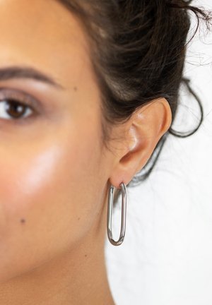 Silver hoop earrings with an elongated design, featuring a smooth metal texture and a polished finish, worn on a person's ear.