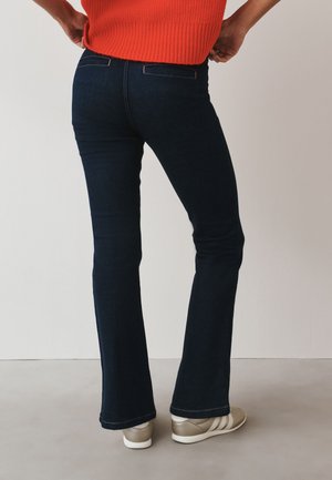 Osoba oblečená v tmavomodrých bootcut džínsoch, béžových teniskách a oranžovom tielku bez rukávov, stojaca s rukami v bokoch, otočená chrbtom.