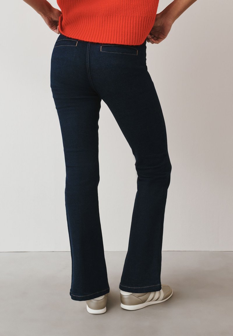Personne portant un jean bootcut bleu foncé, des baskets beiges et un haut sans manches orange, debout, mains sur les hanches, vue de dos.