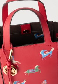 Κόκκινη δερμάτινη τσάντα με πολύχρωμες απεικονίσεις αλόγων, χρυσό κυκλικό μενταγιόν με λογότυπο και διπλές λαβές σε λευκό φόντο.