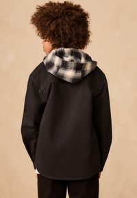 Giacca nera con cappuccio a quadri. Il cappuccio presenta motivi a scacchi bianchi e neri. La giacca ha polsini con bottoni e una vestibilità morbida.