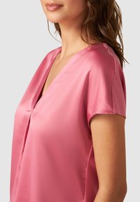 Růžová saténová blůza s výstřihem do V, krátkými rukávy a jemným skladem v přední části. Hladká textura a volný střih.