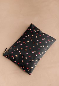 Juodos spalvos kišenė su užtrauktuku, puošta raudonų, rožinių ir oranžinių žvaigždžių raštais, padėta ant kreminės spalvos paviršiaus.