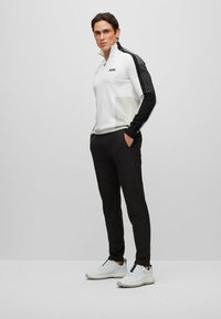 Biela mikina na zips s čiernymi rukávmi a šedým akcentovým vreckom, v kombinácii s čiernymi nohavicami a bielymi športovými topánkami. Minimalistický dizajn.