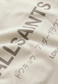 Tissu beige à texture lisse avec un grand texte gris indiquant "ALL SAINTS" ainsi que du texte supplémentaire en japonais et "LONDON".