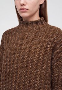 Gros plan d'une personne portant un pull épais en tricot texturé marron avec un col montant et des manches longues.