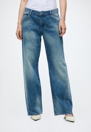 Personne portant un jean bleu à jambes larges et des talons blancs pointus, debout devant un fond clair uni.
