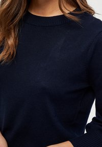Pull en tricot bleu marine avec un col rond et des poignets côtelés, présentant une texture lisse et une coupe ajustée.