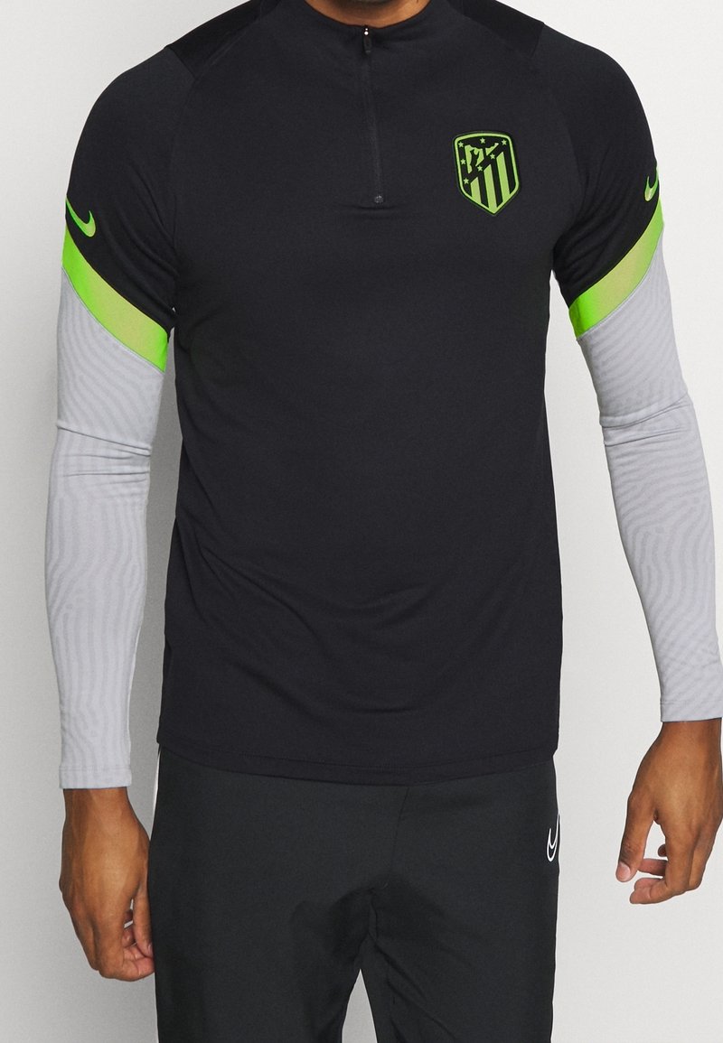 Schwarzes Sportshirt mit halbhohem Reißverschluss, grünen Akzenten an den Ärmeln und einem Logo. Die langen Ärmel haben eine hellgraue gestreifte Struktur.