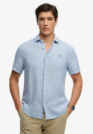 Man draagt een lichtblauw-wit gestreept overhemd met korte mouwen en een beige broek, handen in de zakken tegen een witte achtergrond.