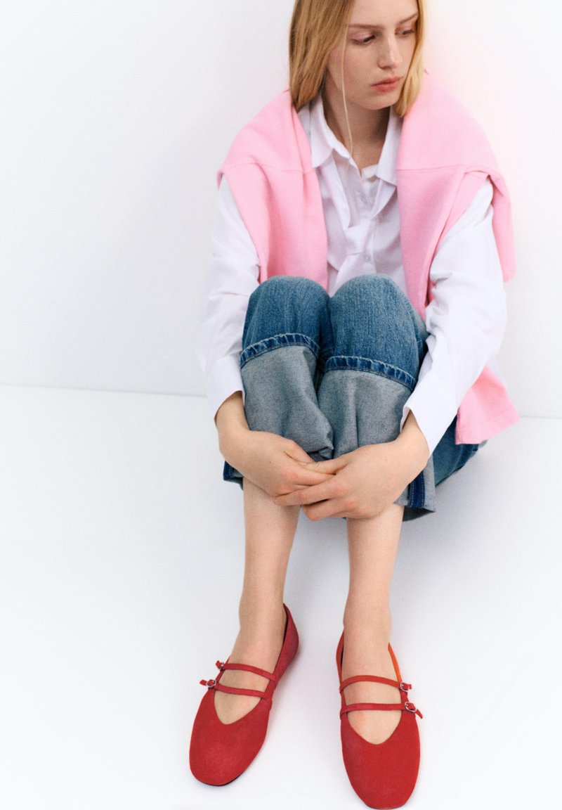 Jeune femme assise par terre, genoux pliés, portant un jean retroussé, une chemise blanche, un pull rose posé sur les épaules et des chaussures rouges Mary Jane.