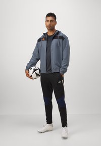 Grå sportsjakke med sorte accenter, sorte og blå bukser, hvid fodbold og hvide fodboldsko; slank, atletisk pasform og casual design.