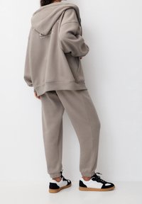 Ljusgrå oversized hoodie med huva och muddar på ärmarna, matchad med byxor i samma färg med muddar och svart-vita sneakers.