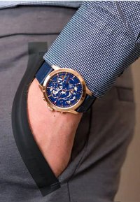 Montre bleue avec un cadran squelette et des accents dorés, dotée d'un bracelet en cuir bleu, reposant sur la poche d'une chemise gris.