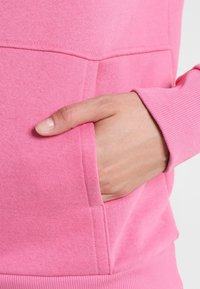 Primer plano de tela de sudadera rosa que presenta un puño acanalado y un bolsillo lateral con una mano descansando dentro, mostrando textura y color.