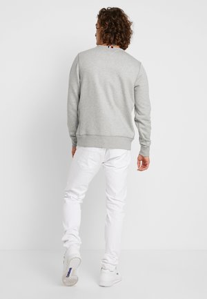 Mann mit lockigem Haar, der einen grauen Pullover, weiße Hose und weiße Sneakers trägt, steht vor einem schlichten hellen Hintergrund und ist von hinten zu sehen.