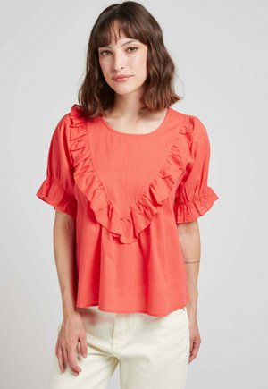 Blouse corail avec des volants le long de l'encolure et des manches, manches courtes bouffantes et coupe décontractée, associée à un pantalon de couleur claire.