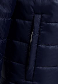 Veste isolante bleu marine avec un design matelassé, une texture lisse et une poche latérale zippée. Le tissu présente un motif en grille subtil.