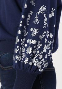 Mörkblå sweatshirt med silverblommönster på ärmarna. Har ribbade muddkragar och en avslappnad passform. Slät textur, avslappnad design.