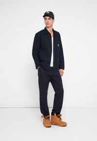 Νεαρός άνδρας που φοράει μαύρο μπουφάν, σκούρο μπλε παντελόνι, καφέ εργασιακά μποτάκια, λευκό πουκάμισο και μαύρο καπέλο μπέιζμπολ με ρίγες, στέκεται μπροστά σε λευκό φόντο.