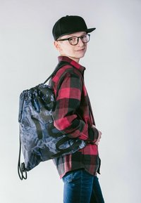 Zaino con coulisse in nero e blu con un motivo grafico; indossato sopra una camicia a scacchi rossa e nera, abbinato a jeans blu.