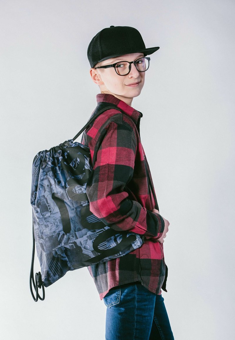 Zaino con coulisse in nero e blu con un motivo grafico; indossato sopra una camicia a scacchi rossa e nera, abbinato a jeans blu.
