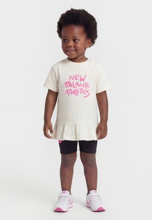 Παιδάκι κοριτσάκι που χαμογελά, φοράει ένα εκρού φόρεμα με ροζ κείμενο "New Balance Athletics", μαύρα σορτς και άσπρα αθλητικά παπούτσια με ροζ λεπτομέρειες.