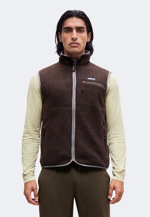 Homme aux cheveux foncés portant une veste polaire marron sur une chemise beige à manches longues, debout contre un fond clair uni.