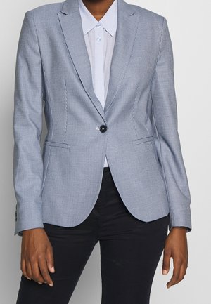 Personne portant un blazer à carreaux bleu clair avec une chemise blanche à col et un pantalon noir, debout devant un fond blanc.