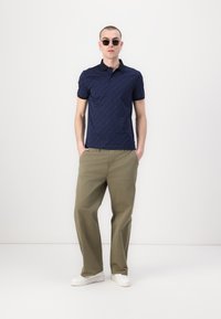 Marineblå polo med teksturert mønster, svart krage, kombinert med løse olivengrønne bukser og hvite sneakers. Modell med solbriller.