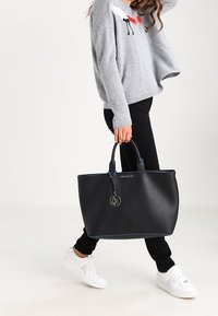 Czarna torba tote z niebieskim akcentem, wykonana z sztucznej skóry, z metką z logo i podwójnymi uchwytami, w połączeniu z codziennym strojem.