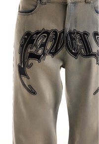 Pantalon marron clair avec un texte brodé de style gothique noir orné sur le haut des cuisses à l'avant, près des poches et de la zone de la fermeture éclair.