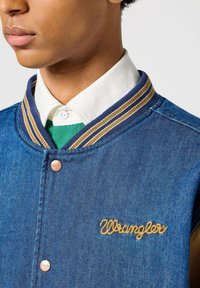 Jeansowa kurtka z zapięciem na guziki, niebieska, z prążkowanym kołnierzem w brązowe i złote paski oraz wyszywanym logo "Wrangler" złotą nicią.