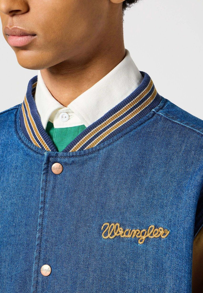 Jeansowa kurtka z zapięciem na guziki, niebieska, z prążkowanym kołnierzem w brązowe i złote paski oraz wyszywanym logo "Wrangler" złotą nicią.