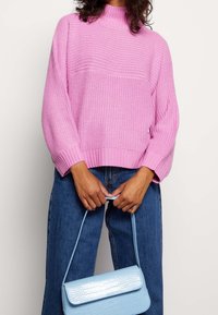 Rosa gestrickter Pullover mit hohem Kragen, langen Ärmeln und einem gerippten Saum, kombiniert mit einer hellblauen Tasche mit Krokodilprägung.