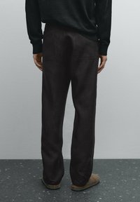 Tummat, rentoistuvaiset housut, jotka on valmistettu kuvioidusta kankaasta ja joissa on kaksi takataskua, nähtynä takaa yksinkertaisen mustan neuleen kanssa.