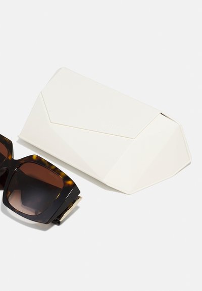 Tortoiseshell sunglasses with dark lenses beside a white, angular, textured case featuring embossed branding.
