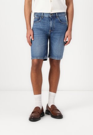 Calções de ganga em azul claro com um corte standard, apresentando cinco bolsos, detalhes de costura e bainha rodada. Sapatos marrons com meias brancas.