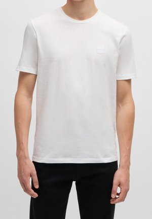 T-shirt en coton blanc à manches courtes, col rond, avec un patch logo texturé sur la poitrine gauche. Porté avec un pantalon noir.