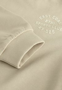 Beige sweatshirt with ribbed cuffs, featuring raised white text "EAST COAST ATHL. SPORTS CLUB MY. 025" and a soft, textured fabric.