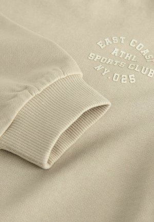 Beige svetr s žebrovanými manžetami, na kterém se nachází vyvýšený bílý text "EAST COAST ATHL. SPORTS CLUB MY. 025" a měkká, texturovaná tkanina.