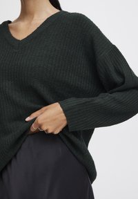 Pull en maille côtelée vert foncé avec un col en V, coupe oversize et manches longues, superposé à une jupe en satin noire. Détails de tissu texturé visibles.